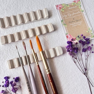 Puede incluir: Tres pinceles de diferentes tamaños descansan en un portapinceles de cerámica blanca. El portapinceles tiene un diseño ondulado y está hecho de cerámica blanca moteada. Los pinceles están hechos de cerdas naturales y tienen mangos de madera. El fondo es una superficie blanca.