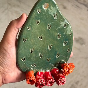May include: A green ceramic wall hanging shaped like a prickly pear cactus pad. The surface features small carved V-shaped indentations and a hole at the top for mounting. Five textured, bright red and orange flower-like protrusions extend from the base.