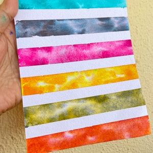 Könnte beinhalten: Ein Aquarellgemälde auf weißem Papier mit horizontalen Streifen in verschiedenen Farben. Die Farben umfassen Türkis, Grau, Rosa, Gelb, Olivgrün und Orange. Das Gemälde wird von einer Hand gehalten.