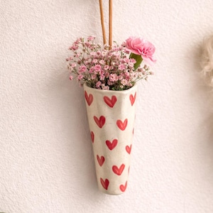 May include: A cream ceramic vase decorated with red hearts, suspended on a wall. It contains pink flowers and a pink carnation. A brown wooden button and a leather cord are used for hanging. The vase is filled with flowers.