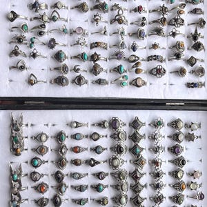 Könnte beinhalten: Eine Sammlung silberfarbener Ringe mit verschiedenen Edelsteinfassungen, darunter Türkis, Amethyst und Onyx. Die Ringe werden in einer schwarzen Schmuckschatulle mit einzelnen Fächern präsentiert und zeigen eine Vielzahl von Designs und Größen.