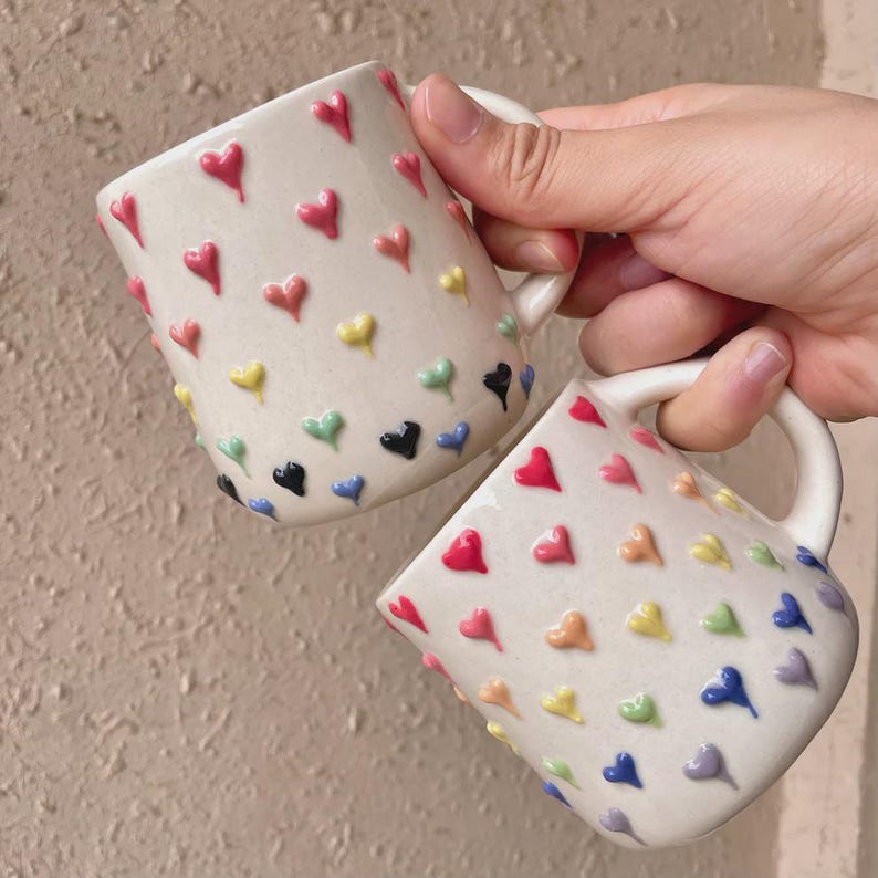 May include: Two white ceramic mugs decorated with raised, colorful heart shapes. The hearts are in a rainbow of colors, including red, pink, yellow, green, blue, and black. Each mug has a white handle.