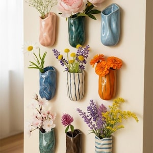 Op de afbeelding: Een verzameling van negen keramische wandvazen in verschillende kleuren, elk met verschillende bloemen. De vazen zijn in tinten roze, blauwgroen, blauw, oranje, bruin en wit met gestreepte en getextureerde ontwerpen. De bloemen zijn onder meer pioenrozen, madeliefjes en lavendel.