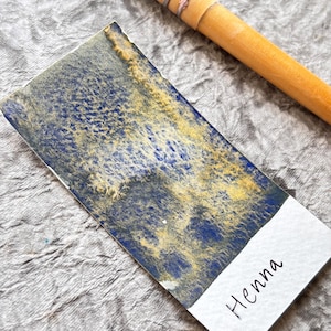 Peut inclure: Un échantillon rectangulaire d'aquarelle avec un motif bleu et or texturé. Le mot "Henna" est écrit à l'encre noire sur une étiquette blanche en bas. Un manche de pinceau en bois est visible en haut à droite.