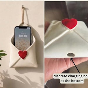 Könnte beinhalten: Ein cremefarbener Keramik-Handyhalter zur Wandmontage in Umschlagform mit einem roten Herzakzent auf der Vorderseite. Der Halter verfügt über eine Juteschnur zum Aufhängen und ein diskretes Ladeloch an der Unterseite für ein Smartphone.