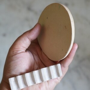 May include: A circular, flat, beige ceramic disc with a smooth surface and a separate rectangular, white ceramic piece featuring a series of vertical, parallel ridges along its length, both held together in a palm-up orientation.