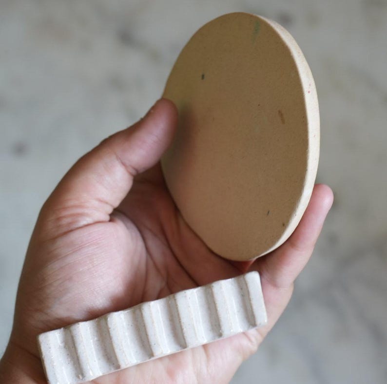 May include: A circular, flat, beige ceramic disc with a smooth surface and a separate rectangular, white ceramic piece featuring a series of vertical, parallel ridges along its length, both held together in a palm-up orientation.
