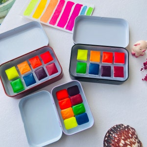 May include: Three open metal tins containing watercolor paint pans. The top tin has a black lid and contains 12 bright, colorful watercolor pans. The middle tin has a brown lid and contains 12 watercolor pans in shades of red, orange, and yellow. The bottom tin has a purple lid and contains 12 watercolor pans in shades of green, blue, and purple.