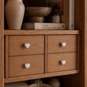 May include: A wooden storage unit featuring four drawers arranged in a two-by-two grid. Each drawer is equipped with a small, white, heart-shaped knob. The unit is constructed from light brown wood with a visible grain, set within a larger shelving unit.