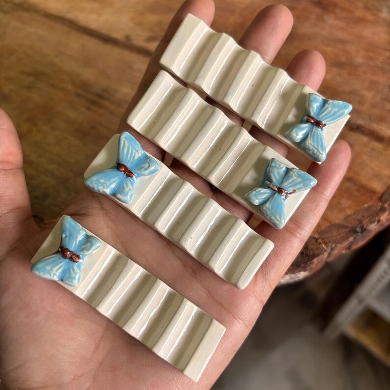 Puede incluir: Juego de cuatro reposa palillos de cer&aacute;mica. Cada reposa palillos es de color blanco roto con un dise&ntilde;o acanalado y un extremo cuadrado con una mariposa azul claro con detalles marrones. Los reposa palillos se sostienen en una mano.