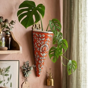 Op de afbeelding: Een levendige oranje wandplanter met een wit bloemmotief, met een groene Monstera-plant. De planter is kegelvormig en aan een roze muur bevestigd. Een kleine messing verstuiver is ook zichtbaar.