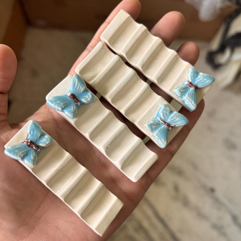Puede incluir: Juego de cuatro reposa palillos de cer&aacute;mica blanquecina. Cada reposa palillos presenta una base rectangular con superficie acanalada y una mariposa azul claro con detalles marrones. Un complemento decorativo para cualquier mesa.