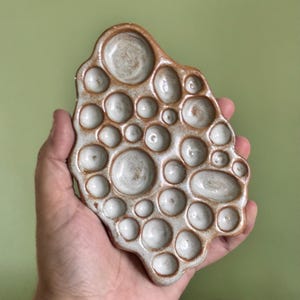 May include: An irregular, organic-shaped ceramic tray featuring a textured, light-colored surface with numerous circular indentations of varying sizes. The edges and the raised areas between the depressions are finished in a warm, earthy brown glaze.