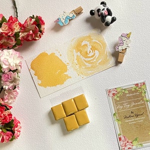 May include: A set of five gold watercolour paint pans, arranged in a T shape. The pans are on a white surface with a watercolour painting of a gold swirl and a card that says "My Tiny Journal" by "Meghana Agarwal" Handmade Watercolour Journals & Paints. Made in India.