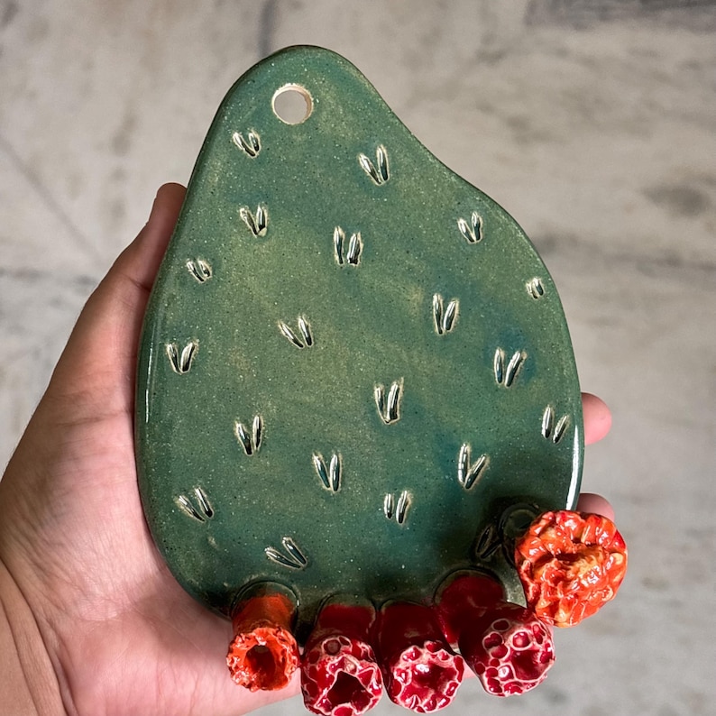 May include: A green ceramic wall hanging shaped like a prickly pear cactus pad. The surface features small, carved indentations and a circular hole at the top for mounting. Five textured, bright red, cylindrical flower-like protrusions are attached.