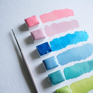 May include: A collection of watercolor paints in various shades of pink, blue, and green, arranged with corresponding brush strokes on white paper. A thin, light-colored paintbrush is positioned near the paints, suggesting artistic use. The paints are in small, square containers.