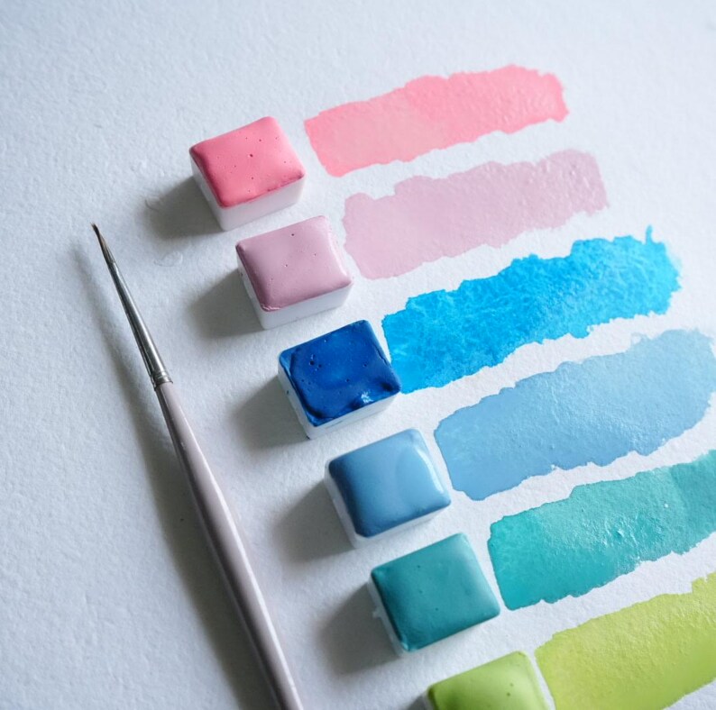 May include: A collection of watercolor paints in various shades of pink, blue, and green, arranged with corresponding brush strokes on white paper. A thin, light-colored paintbrush is positioned near the paints, suggesting artistic use. The paints are in small, square containers.