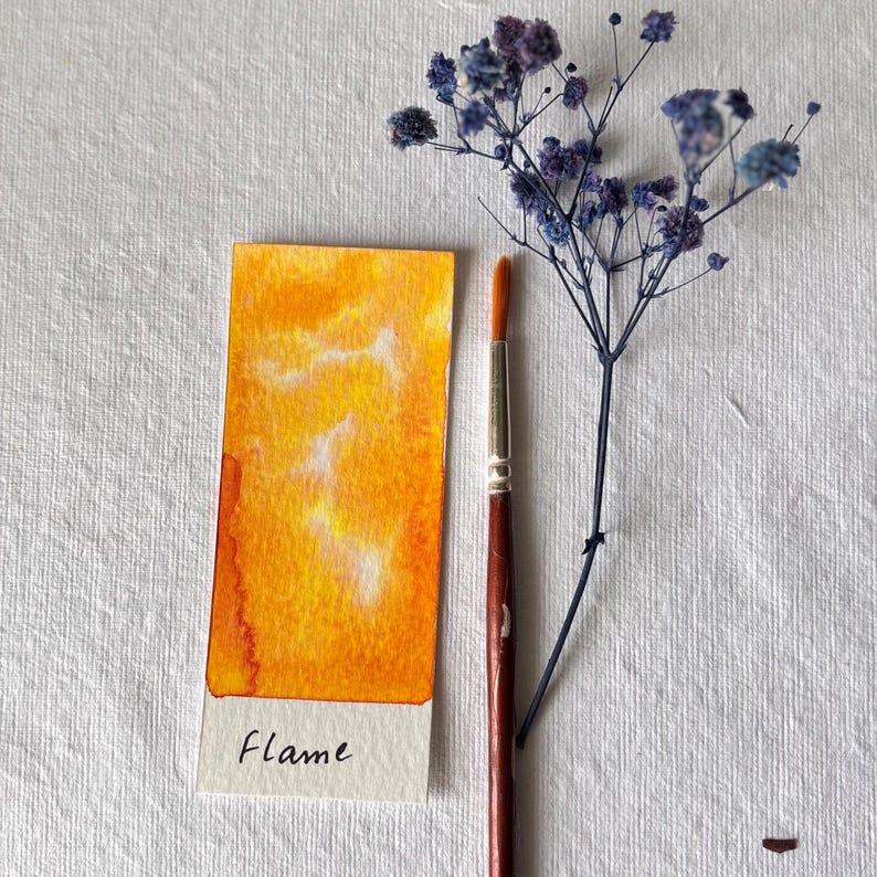 K&ouml;nnte beinhalten: Ein rechteckiges Aquarellgem&auml;lde mit gelben und orangen Farbt&ouml;nen, betitelt "Flame". Ein Pinsel und ein Zweig getrockneter blauer Blumen liegen neben dem Gem&auml;lde auf einer wei&szlig;en, strukturierten Oberfl&auml;che.