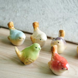 May include: A selection of ceramic bird ornaments in shades of green, red, and blue, with orange and yellow detailing. The birds are displayed on a light-coloured wooden surface, highlighting their intricate design and glossy finish.