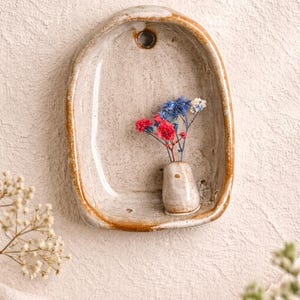 Può includere: Una tasca da parete in ceramica bianco sporco fatta a mano con un piccolo vaso contenente fiori secchi rossi, blu e bianchi. La tasca ha una forma rustica e irregolare con un bordo marrone e un foro per appenderla.