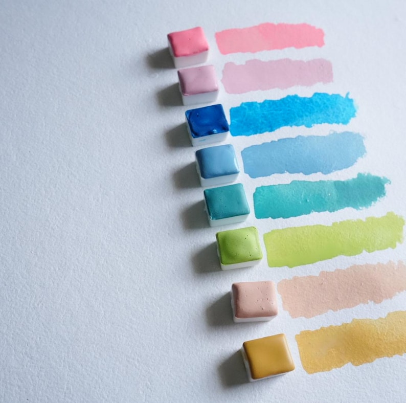 May include: A collection of watercolor paint squares in various colors, including pink, blue, green, and yellow, with corresponding paint swatches. The paint is arranged on a white surface, showcasing the range of hues and tones available for artistic use.