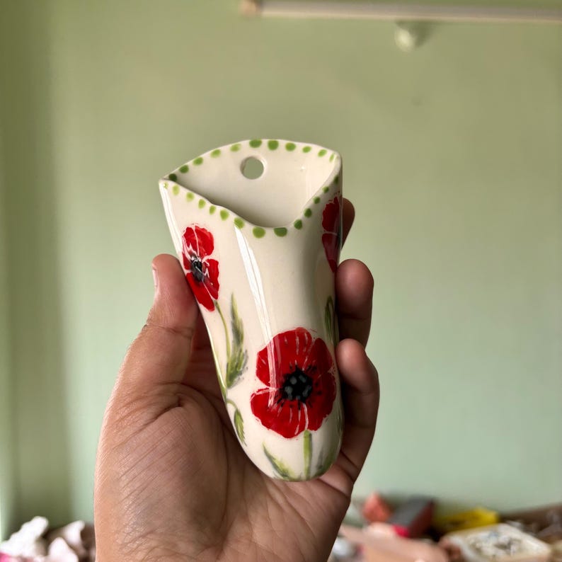 Może przedstawiać: Kremowy ceramiczny wazon ścienny z ręcznie malowanymi czerwonymi makami z czarnymi środkami i zielonymi łodygami. Krawędź ozdobiona jest serią małych zielonych kropek i posiada pojedynczy okrągły otw&oacute;r do zawieszenia na ścianie.