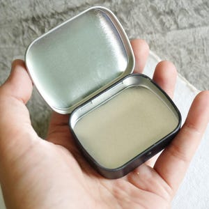 May include: An open, small, rectangular tin with a hinged lid. The tin is held in a hand, revealing the interior. The tin's interior and lid are silver, while the exterior is black. The lid is open, showing the inside of the tin.
