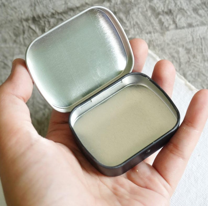 May include: An open, small, rectangular tin with a hinged lid. The tin is held in a hand, revealing the interior. The tin's interior and lid are silver, while the exterior is black. The lid is open, showing the inside of the tin.