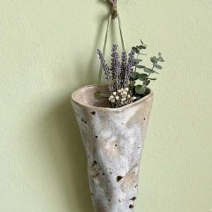 Puede incluir: Una maceta de pared de cerámica en forma de cono con un esmalte moteado en blanco y bronceado, suspendida por una cinta verde claro de un gancho de color bronce con forma de rama. La maceta contiene un pequeño arreglo de lavanda y eucalipto.