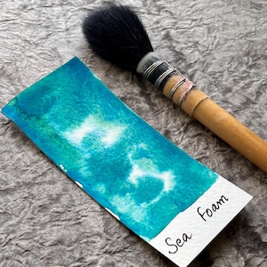 May include: A watercolour painting on a white rectangular paper with the text "Sea Foam". The painting features shades of turquoise and white, resembling sea foam. A large black-bristled brush with a wooden handle rests beside the painting.
