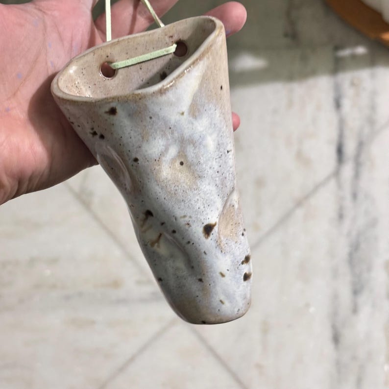 May include: A cone-shaped ceramic wall planter with a speckled beige and white glaze. The vessel features two circular holes at the top rim for hanging, threaded with a light-colored cord. The surface has a smooth, glossy finish with dark spots.