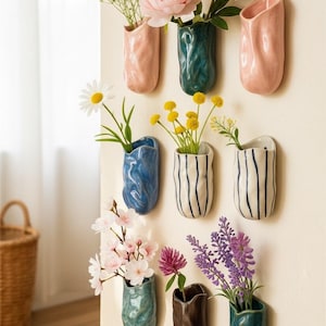 May include: A selection of ceramic wall vases in shades of pink, teal, blue, and brown. Each vase displays different flowers, including pink peonies, daisies, and lavender. The vases are mounted on a cream-coloured wall.