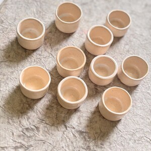 May include: Ten small, white ceramic cups with a slightly textured surface. The cups are arranged in a semi-circle on a light gray textured surface.