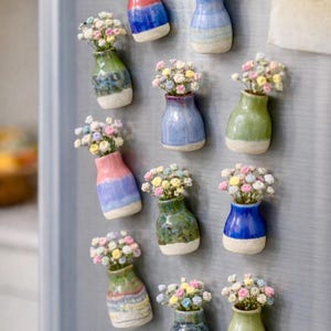May include: A display of miniature ceramic vases, each holding a small bouquet of pastel-coloured flowers. The vases, in shades of blue, green, and pink, are attached to a metallic surface, possibly a fridge. A delightful home decoration.