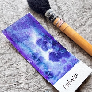 Può includere: Un dipinto ad acquerello nei toni del blu e del viola su un foglio di carta rettangolare, con la parola "Cobalto" scritta in basso. Un pennello con manico in legno e setole nere è appoggiato accanto.