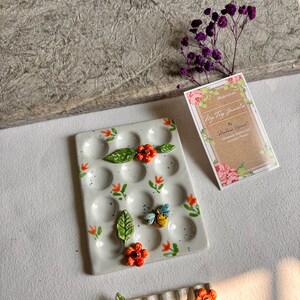 May include: A white ceramic palette with 18 wells, decorated with orange flowers, green leaves, and a bee. The palette is surrounded by a white background.