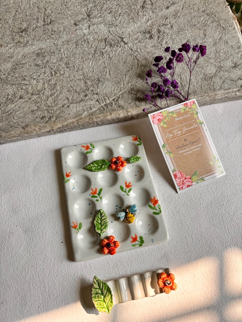 May include: A white ceramic palette with 18 wells, decorated with orange flowers, green leaves, and a bee. The palette is surrounded by a white background.