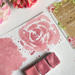May include: A watercolor painting of a pink rose on a white paper background. The painting is surrounded by four small pans of pink watercolor paint.