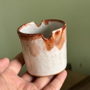 May include: A handmade ceramic ashtray with a speckled white and brown glaze. The rim and upper portion of the ashtray are a rich brown colour, while the lower part is white with brown speckles. The ashtray has a unique, organic shape.