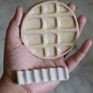 May include: A round, beige ceramic paint palette featuring a grid of twelve recessed square wells for mixing colors, paired with a separate, rectangular, off-white ceramic brush rest that has six parallel grooves along its top surface.
