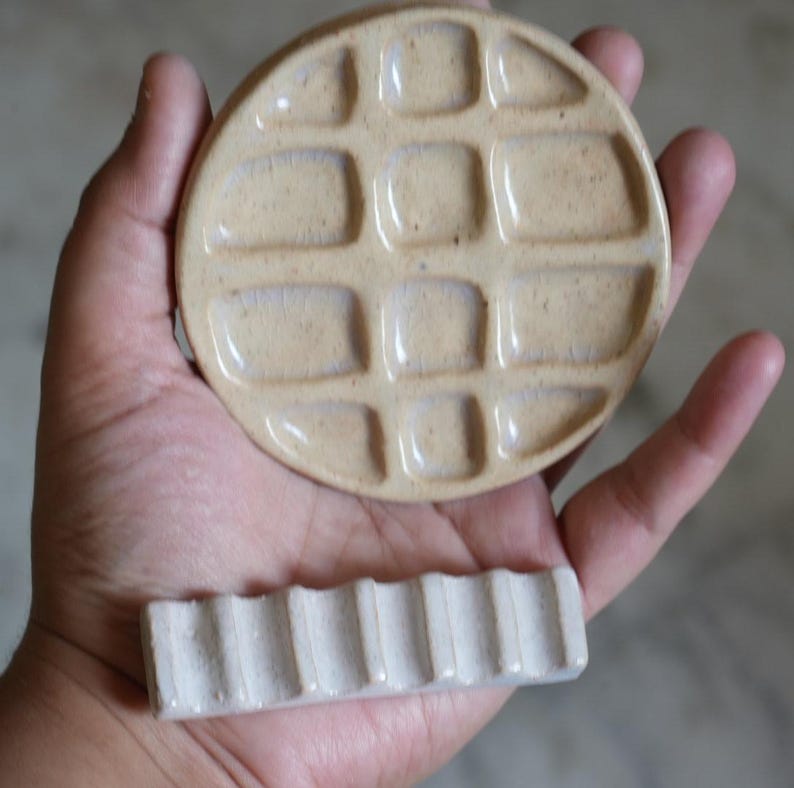 May include: A round, beige ceramic paint palette featuring a grid of twelve recessed square wells for mixing colors, paired with a separate, rectangular, off-white ceramic brush rest that has six parallel grooves along its top surface.