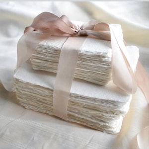 May include: Two stacks of handmade paper, each with a deckled edge, are tied together with a wide, light brown satin ribbon. The paper is a soft white colour, and the ribbon is tied in a bow.