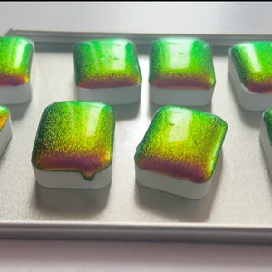 K&ouml;nnte beinhalten: Acht schimmernde Aquarellfarben in einer Metallbox. Die Farben wechseln von Gr&uuml;n zu Gold und Rosa. Jede rechteckige Pfanne hat eine wei&szlig;e Basis. Ideal f&uuml;r K&uuml;nstler.