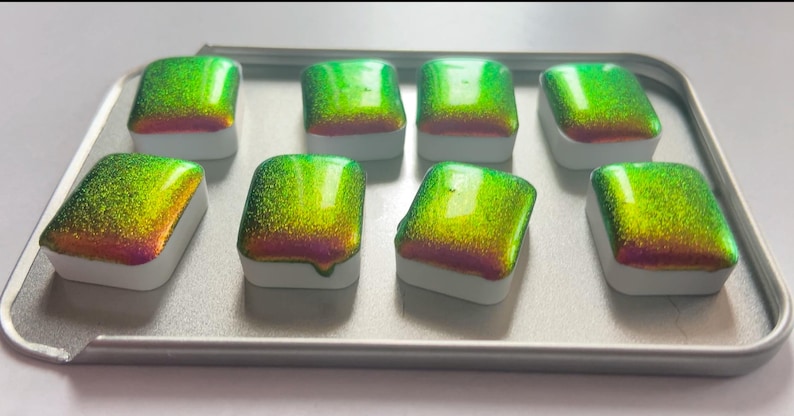 K&ouml;nnte beinhalten: Acht schimmernde Aquarellfarben in einer Metallbox. Die Farben wechseln von Gr&uuml;n zu Gold und Rosa. Jede rechteckige Pfanne hat eine wei&szlig;e Basis. Ideal f&uuml;r K&uuml;nstler.