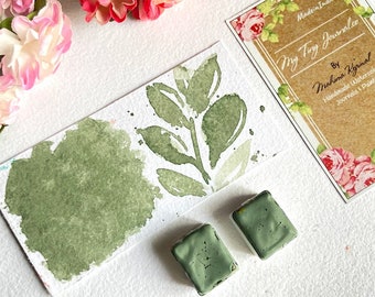 May include: Two green watercolor paint pans with a watercolor painting of a green leaf branch on a white background. The painting is next to a card that says "My Tiny Journal" by "Mahima Kgrinal" Handmade Watercolor Journals & Paint.