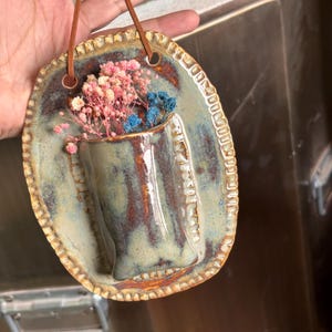Könnte beinhalten: Eine handgefertigte Keramik-Wandtasche mit eingebauter Vase, glasiert in Blau-, Braun- und Grüntönen. Die Vase enthält rosa und blaue Trockenblumen. Zum Aufhängen ist eine braune Lederschnur befestigt.
