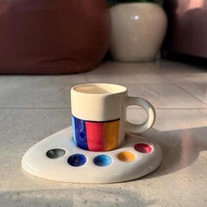 Puede incluir: Una pequeña taza de café de cerámica blanca con una banda colorida de azul, rojo y amarillo. La taza se encuentra sobre un platillo blanco a juego con forma de paleta de artista con pozos de pintura de colores. El diseño es moderno y artístico.