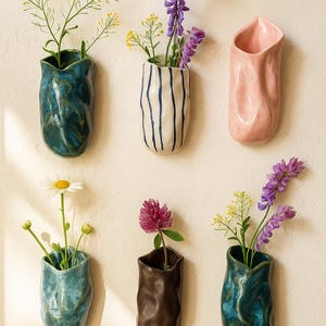 Könnte beinhalten: Sechs keramische Wandvasen in verschiedenen Farben, jede mit frischen Blumen. Die Vasen sind in Blautönen, Weiß mit blauen Streifen und Rosa gehalten und haben ein strukturiertes, organisches Design. Die Blumen umfassen gelbe und violette Blüten.
