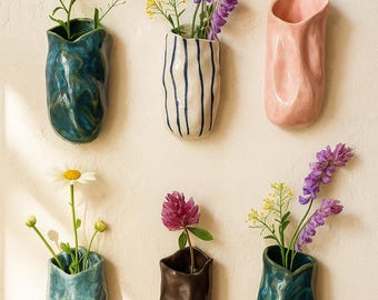 Puede incluir: Seis jarrones de cerámica para pared con formas orgánicas irregulares. La colección incluye un jarrón azul oscuro, uno blanco con rayas azules, uno rosa sólido, uno azul claro, uno marrón oscuro y uno verde azulado, algunos con flores amarillas y moradas.