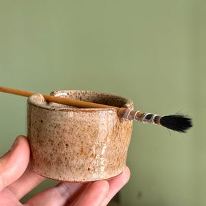 Könnte beinhalten: Kleiner, handgefertigter Keramik-Pinselhalter mit gesprenkelter, hellbrauner Glasur. Ein Pinsel mit Holzgriff und schwarzen Borsten liegt in der Halterung. Der Halter wird in einer Hand vor einem weichen grünen Hintergrund gehalten.